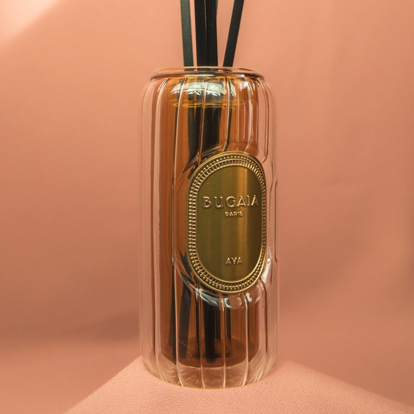 Elegantiškas BUGAIA namų kvapas "Ava" stikliniame buteliuke su medinėmis lazdelėmis, 200 ml, pastatytas ant modernaus paviršiaus. Gėlių ir rytietiškų natų įkvėptas aromatas perteikia prabangą ir jausmingumą. Fone subtilus interjero dekoro elementas.