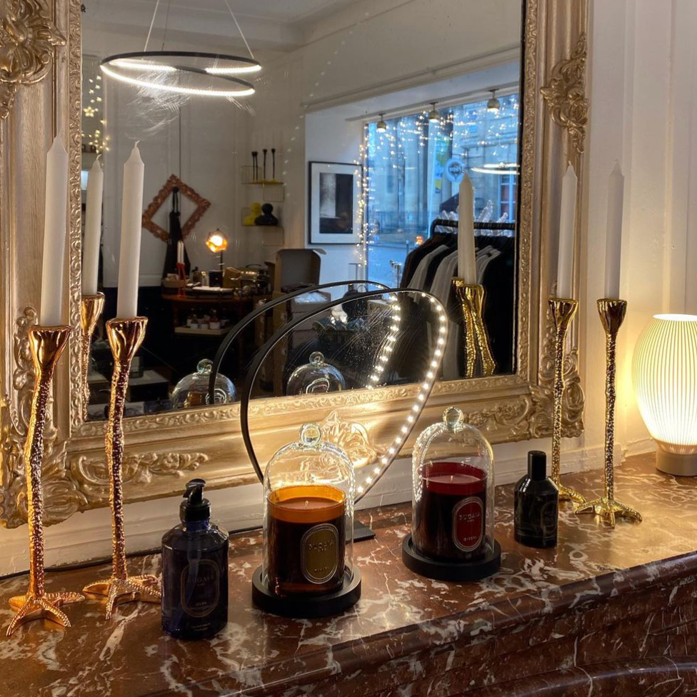 Stilingas BUGAIA stiklinis gaubtas kvapioms žvakėms – skaidrus, elegantiškas kupolas pastatytas ant paviršiaus šalia žvakės. Prabangus interjero akcentas, sukurtas kvapo ir estetikos išsaugojimui. Fone – jaukus namų ar studijos interjeras.