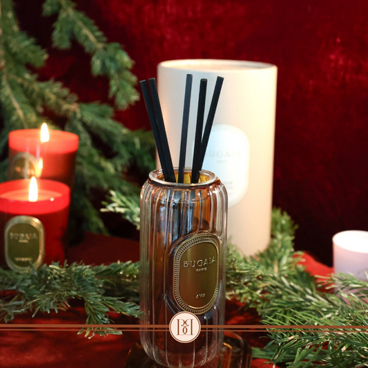 Elegantiškas BUGAIA namų kvapas "Ava" stikliniame buteliuke su medinėmis lazdelėmis, 200 ml, pastatytas ant modernaus paviršiaus. Gėlių ir rytietiškų natų įkvėptas aromatas perteikia prabangą ir jausmingumą. Fone subtilus interjero dekoro elementas.